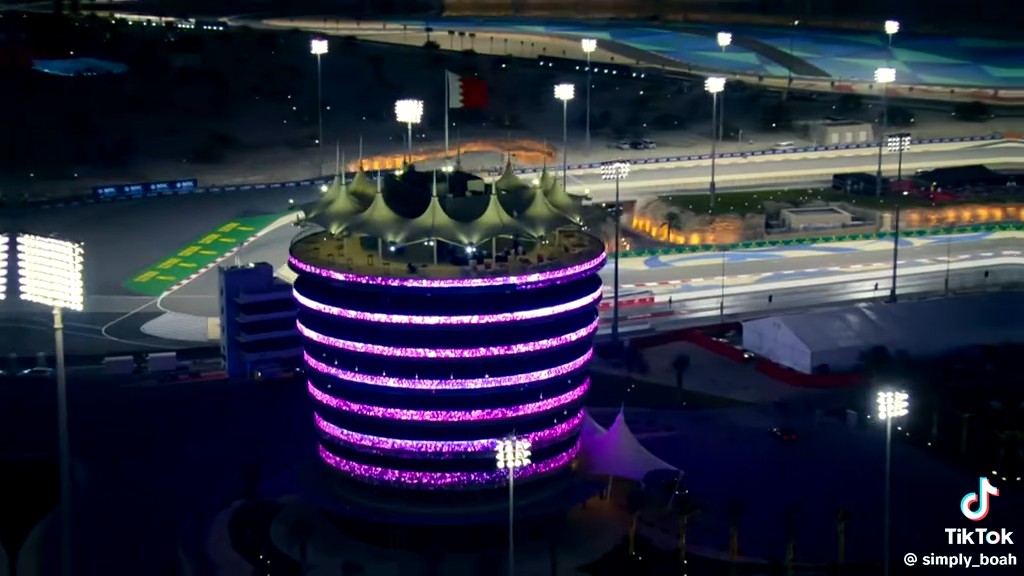 An aerial nighttime view of an illuminated cylindrical tower with purple LED lighting at what appears to be the Bahrain International Circuit, marked with a TikTok watermark '@simply_boah'.