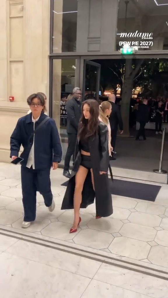 A nighttime street scene outside Madame Figaro showing a woman in a long black leather coat and short black outfit walking with a companion, with text overlay reading '(PFW P/E 2027)'.