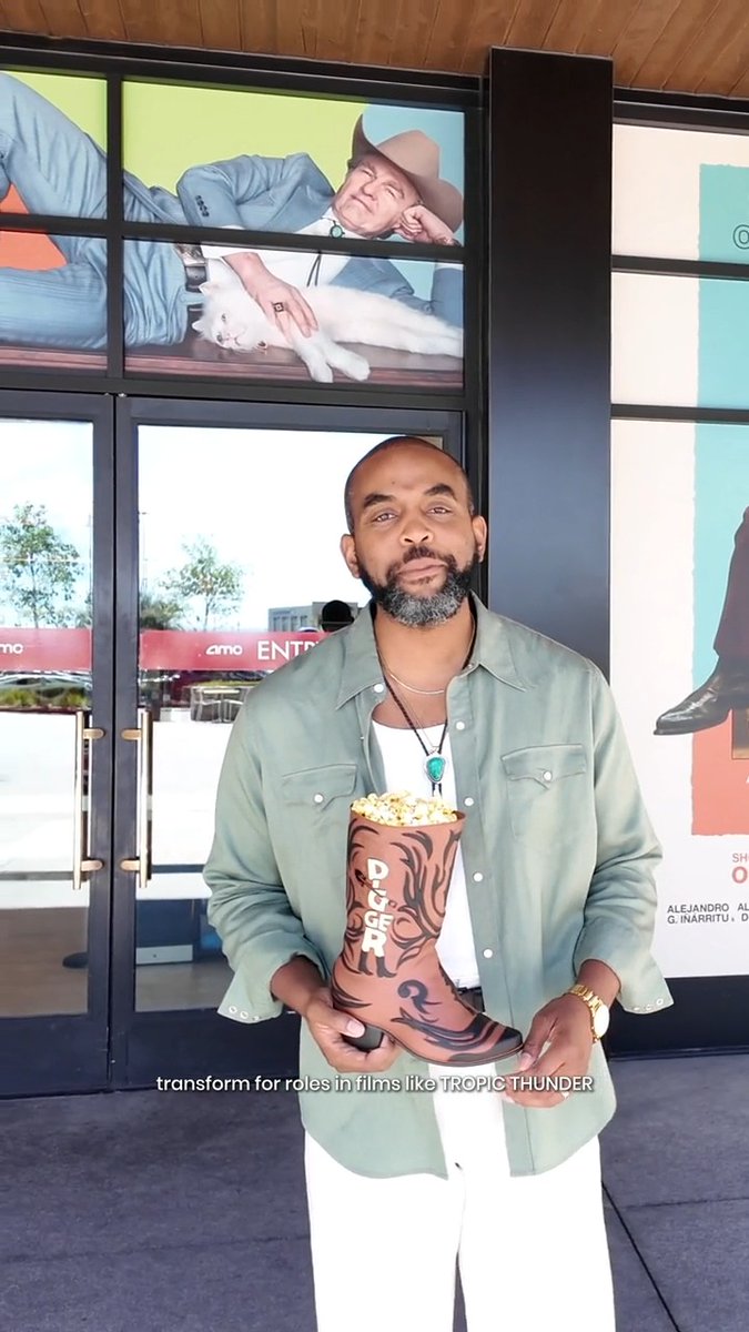 A man stands outside an AMC theater holding a Digger boot-shaped popcorn container, with a screen behind him advertising "Alejandro G. Iñárritu" as director, promoting the new film tie-in to Tom Cruise's comedy.