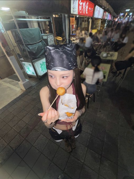 A young woman wearing a black bandana and crop top eats a small round fried food on a stick while standing in a busy night market.