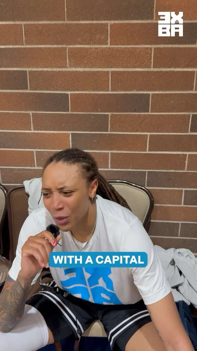 WNBA player Brittney Griner, wearing a white t-shirt and athletic shorts, sits in front of a brick wall with a distressed facial expression while holding a small microphone; text on screen reads 'WITH A CAPITAL' and the 'EX BR' logo appears in the corner.