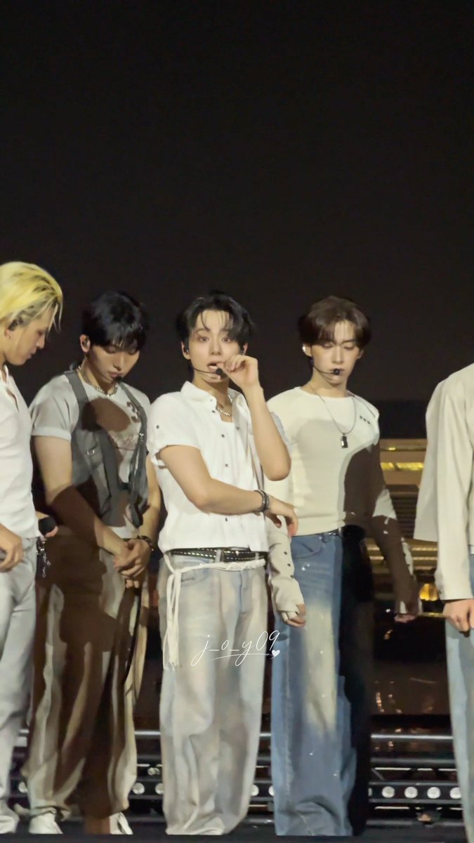 Four male K-pop performers stand on a dimly lit stage wearing coordinated outfits of white tops and light jeans, with a watermark 'j_o_y09' visible across the center.