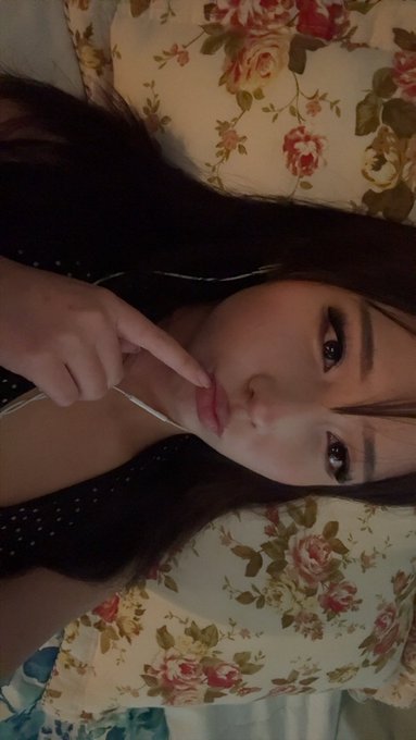A close-up selfie of a young woman lying on a floral pillow, with her index finger pointing toward her lips.