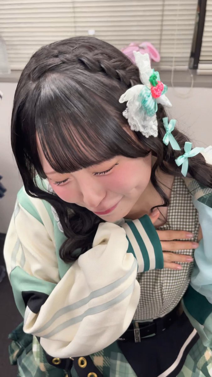 A young woman with braided hair and decorative hair clips, including a strawberry, smiles gently while wearing a green and white outfit.