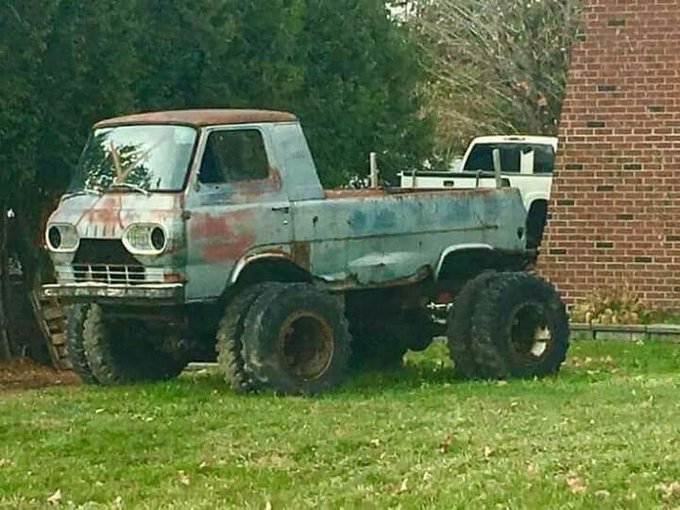 An old, rusted, vintage pickup truck with oversized tires parked on grass next to a brick building.