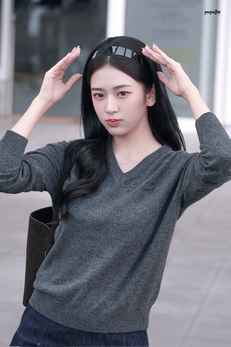A young woman with long dark hair wearing a gray V-neck sweater and jeans, posing with both hands raised near her head outdoors.