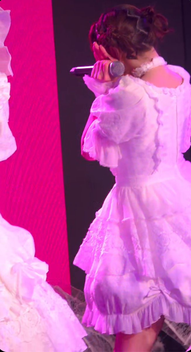 A performer on stage is captured from behind wearing an elaborate, ruffled pink and white frilly dress, holding a microphone under bright pink stage lighting.