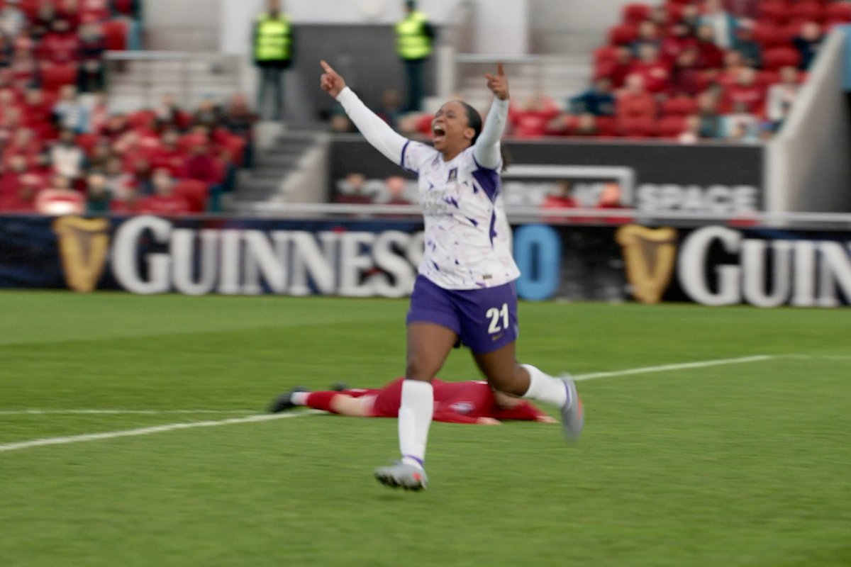 A soccer player wearing jersey number 21 in a white and purple kit celebrates jubilantly with both arms raised, as an opponent lies on the ground behind him, with "GUINNESS" visible on the stadium advertising boards. The image captures a triumphant moment that embodies the "Progress is everything" spirit of #TedLasso, celebrating determination and victory on the pitch.