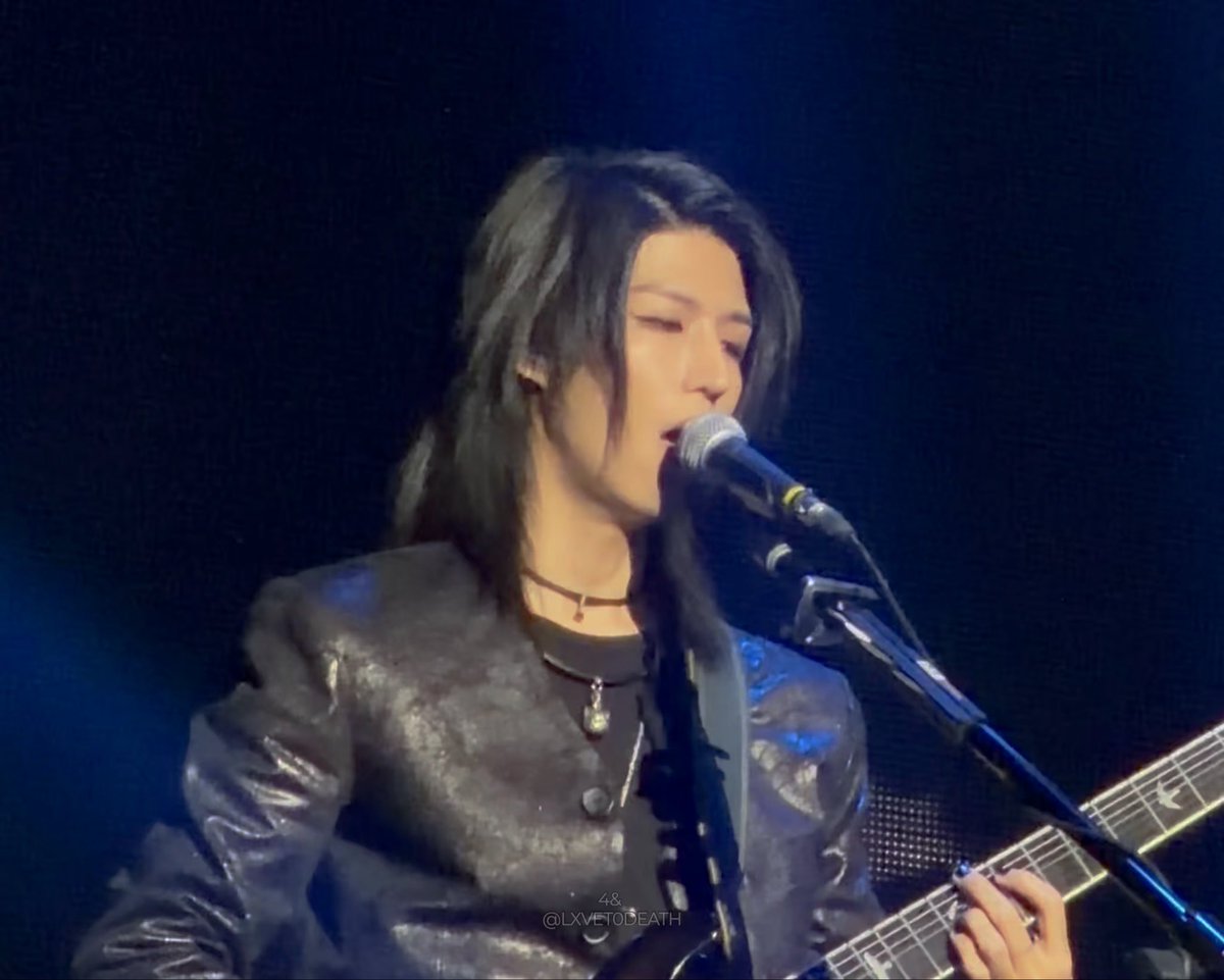 A musician with shoulder-length black hair performing on stage, singing into a microphone while playing a guitar, wearing a shiny silver jacket.