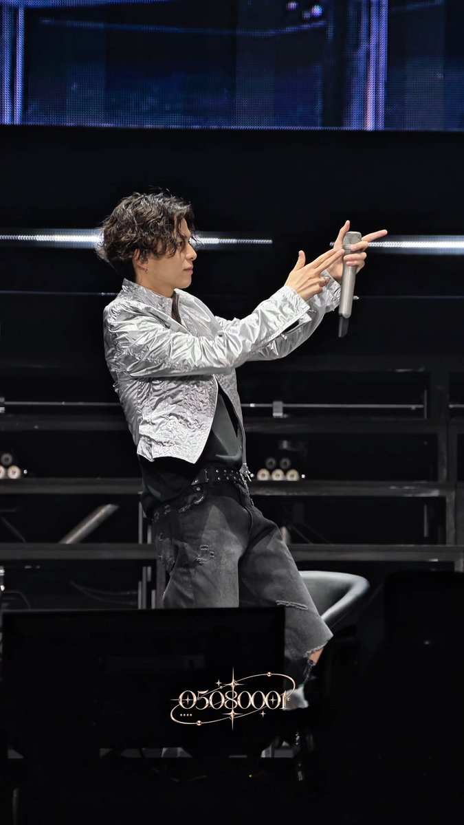 A male performer on stage wearing a metallic silver shirt and black pants, holding a microphone with both hands; the image is signed '051360'.