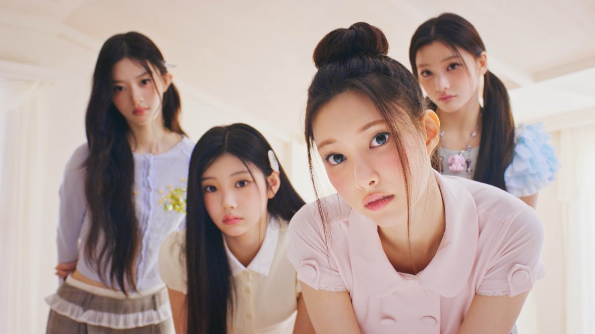 Four young women in pastel-colored outfits pose together indoors, with the girl in the pink top leaning forward closest to the camera.