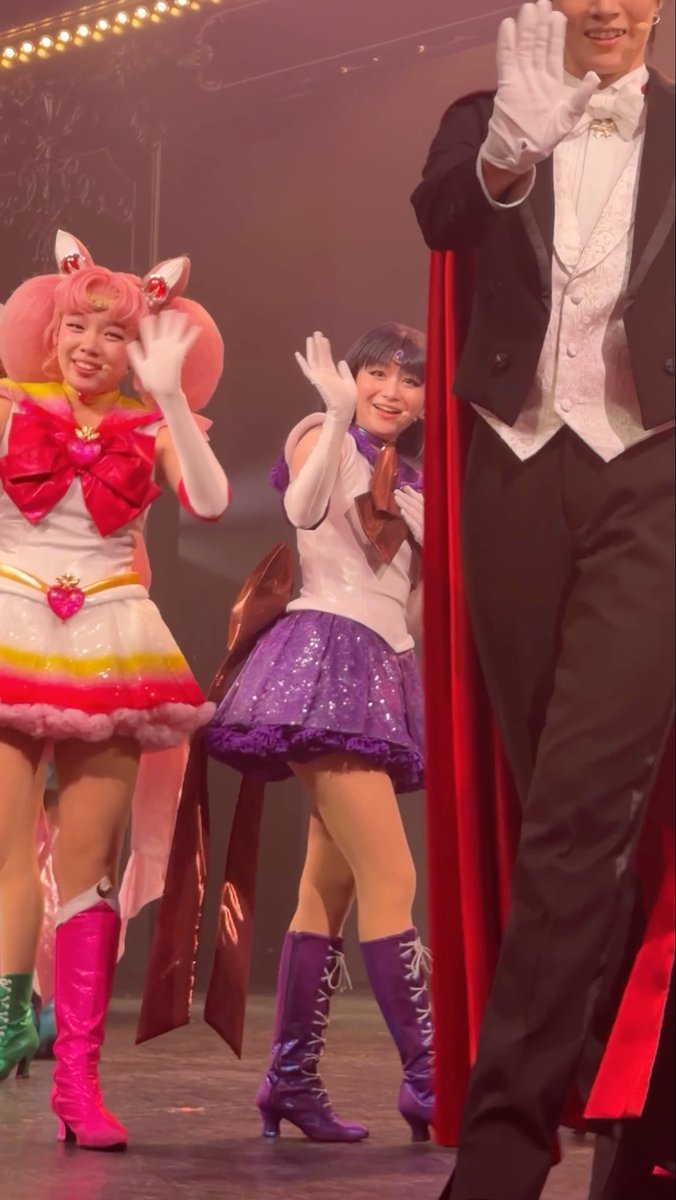 Two performers in Sailor Moon-inspired costumes—one in pink with a large bow and the other in purple with a sparkling skirt and purple boots—waving on stage alongside a person in a tuxedo and white gloves.