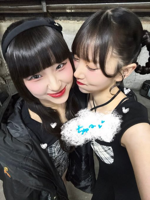 Two young women pose closely together for a selfie; both wear black outfits with white heart details and have dark hair with bangs, one with twin braids.