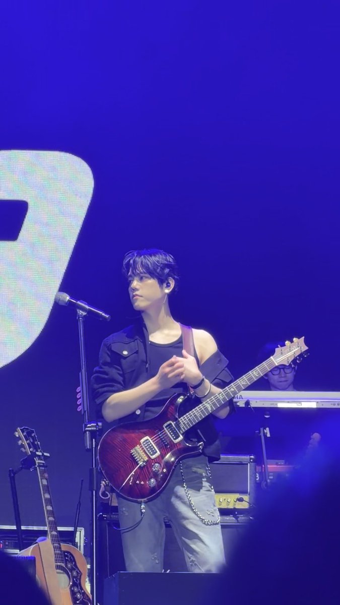 A young male musician with dark hair performs on stage under blue lighting, holding an electric guitar and standing at a microphone stand, with another band member visible at a keyboard in the background.