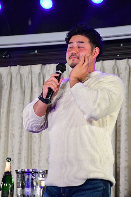 A man in a cream-colored sweater stands on stage holding a microphone close to his face with one hand while touching his cheek with the other, his expression suggesting disbelief—matching the post's sentiment of "No way." Two bottles and a container sit on a table beside him, with stage lights and curtains visible in the background.