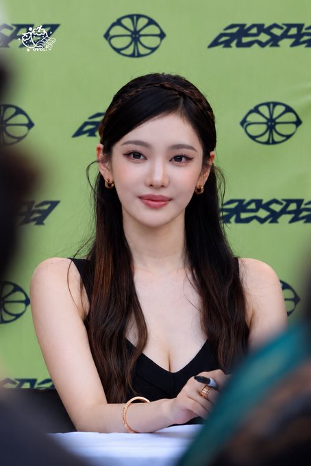 A young woman with long dark hair and a black top poses for a photo against a green backdrop featuring a repeating dark logo pattern.