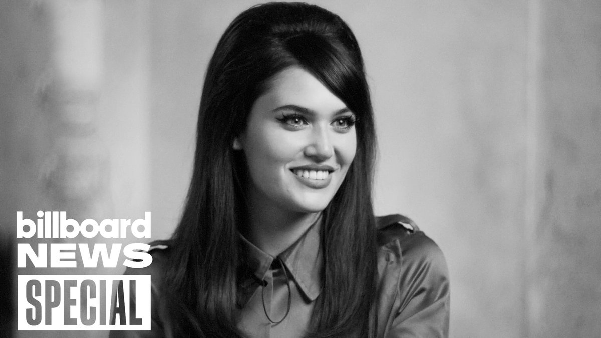 A black-and-white image shows a young woman with long, dark, voluminous hair styled in a 1960s-inspired bouffant, smiling at the camera while wearing a satin button-up shirt. The image features "Billboard NEWS SPECIAL" text in the lower-left corner, complementing the accompanying post about Sienna Spiro discussing her 1960s-inspired look and how it helped her overcome insecurities.