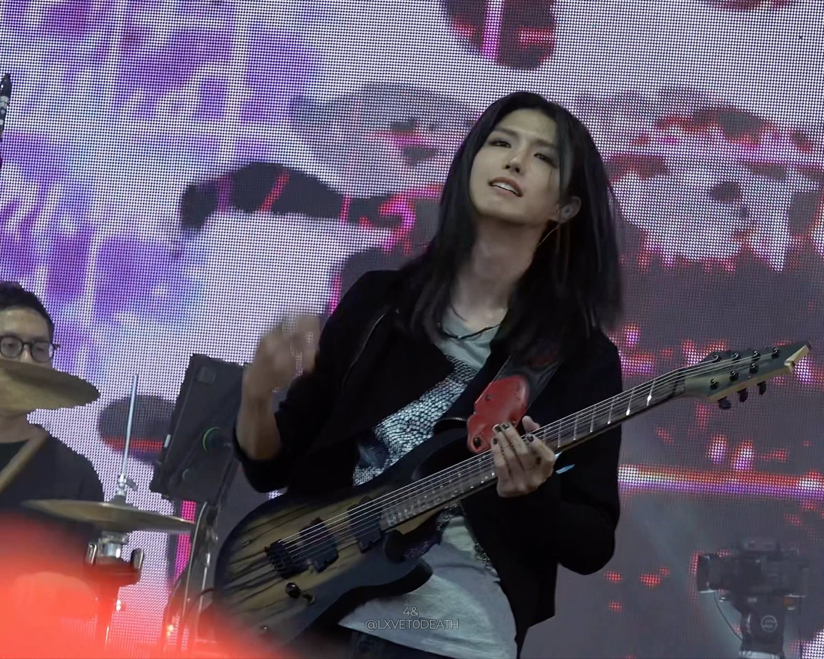 A musician with long dark hair plays an electric guitar on stage, with a drummer visible on the left and a watermark reading '48 @LXVT0DEATH'.