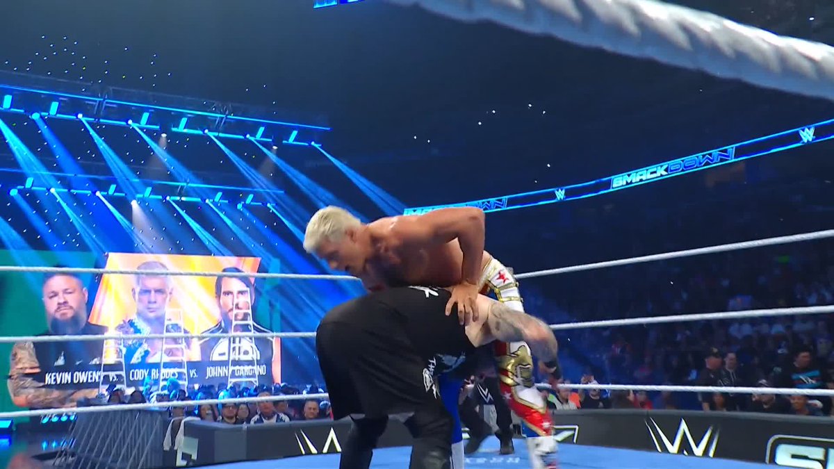 Cody Rhodes is seen fighting in the ring, appearing to struggle against a larger opponent with platinum blonde hair. A SmackDown graphic in the background advertises an upcoming match between Kevin Owens, Cody Rhodes, and Johnny Gargano, hinting at the chaotic brawl that led to this moment.