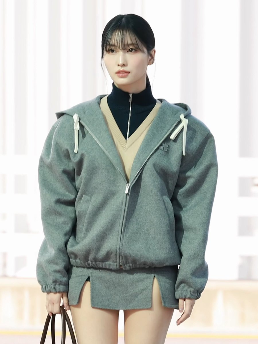 A woman with dark hair and bangs wearing an oversized green fleece hooded jacket layered over a black zip-up top and tan V-neck sweater.