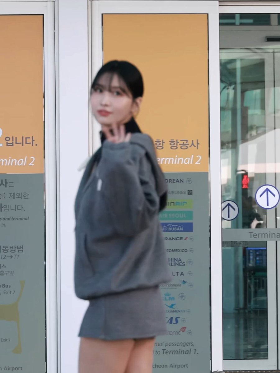 A young woman in a gray hoodie stands at what appears to be an airport Terminal 2 entrance, posing near signage in Korean and English that lists various airlines like Air Seoul, Air Busan, and others.