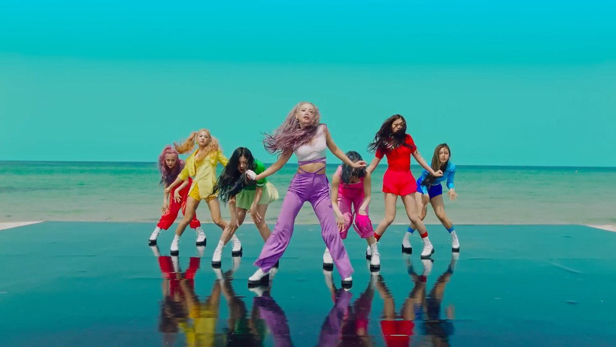 A group of female performers in colorful outfits dance on a reflective turquoise surface against a beach and ocean backdrop.