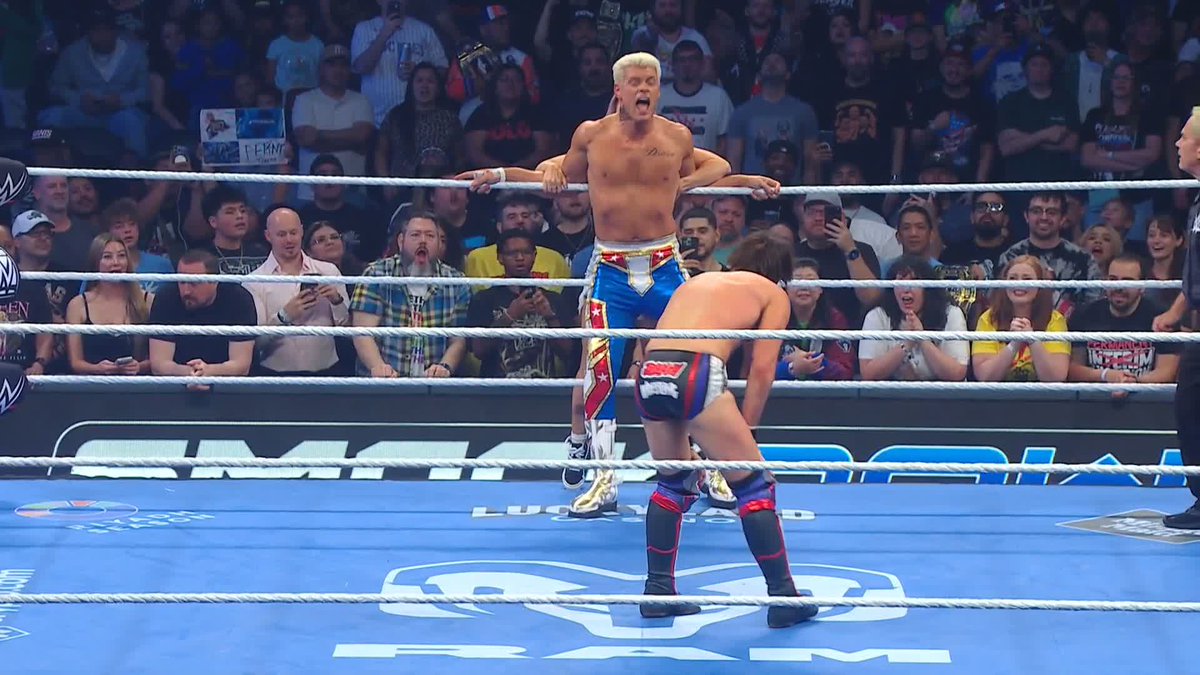 Cody Rhodes, wearing patriotic red, white, and blue ring gear, leans over the ropes in the ring during a WWE SmackDown event, with the ring apron displaying "WWE" and "PRIME" branding. The image captures the moment referenced in the caption, where Cody improvised by whispering to opponent Johnny Gargano to re-execute a table-breaking spot after it failed initially.
