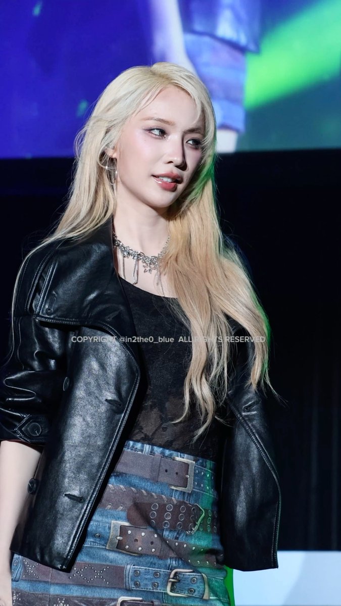 A woman with long blonde hair wears a black leather jacket over a black top, a layered silver necklace, and a blue denim skirt with multiple brown belts; the photo bears the watermark 'COPYRIGHT @in2the0_blue ALL RIGHTS RESERVED'.