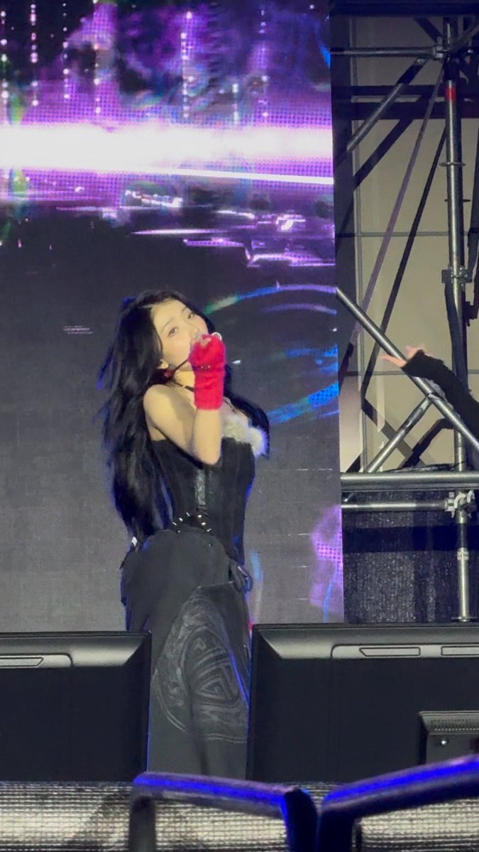 A female performer with long dark hair, wearing a black corset, black pants, and red gloves, sings on stage while holding a microphone in front of a large LED screen.