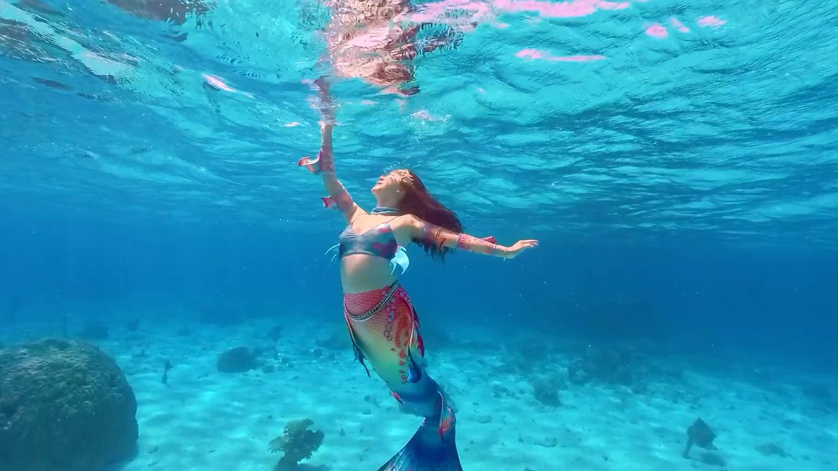 An underwater photograph showing a person wearing a mermaid costume reaching up towards the surface of clear, sunlit tropical water.