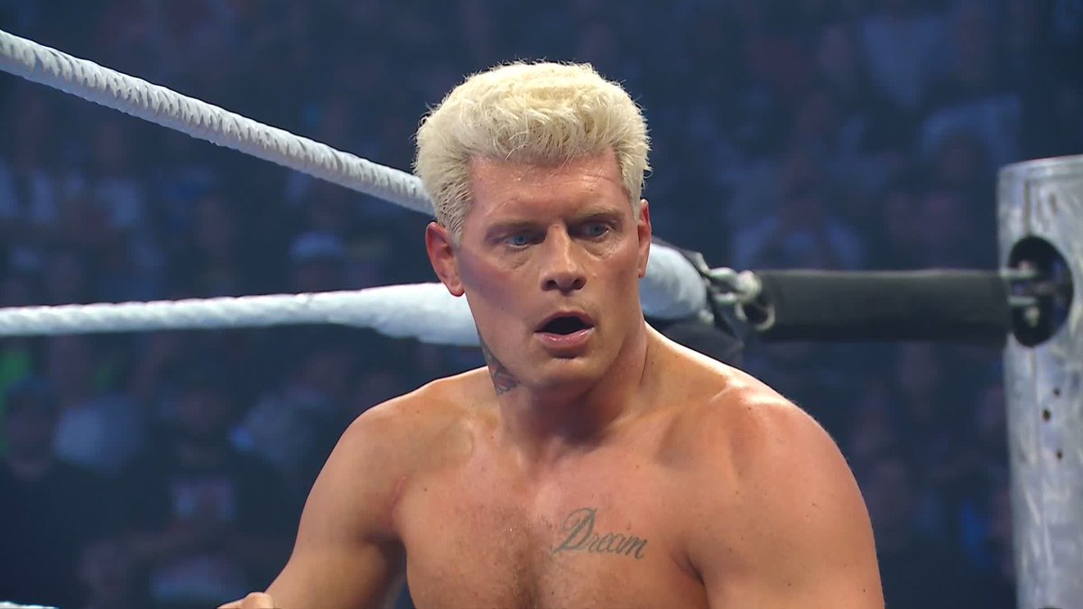 A wrestler with platinum blonde hair stands in the ring with a shocked expression, sporting a "Dream" tattoo on his chest. The caption connects to the post about Johnny Gargano possibly ambushing Cody Rhodes, with the wrestler appearing caught mid-reaction in the ring.