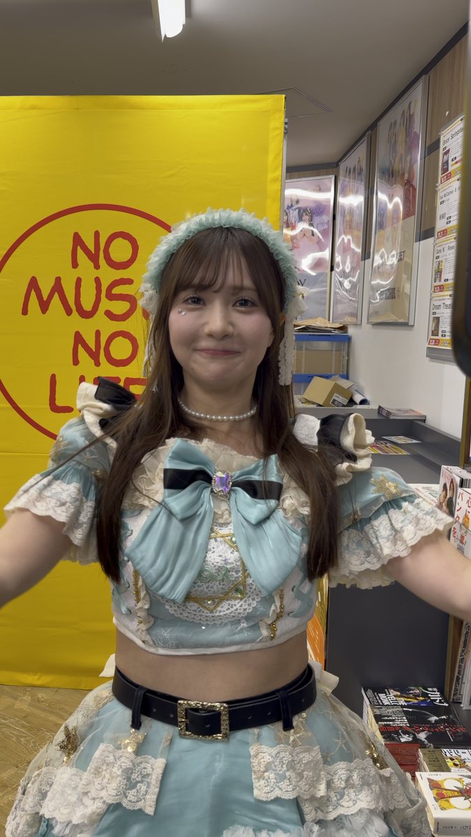 A young woman in a light blue and white frilly idol costume and pearl necklace poses in front of a yellow banner that reads 'NO MUSIC NO LIFE' in what appears to be a record store.