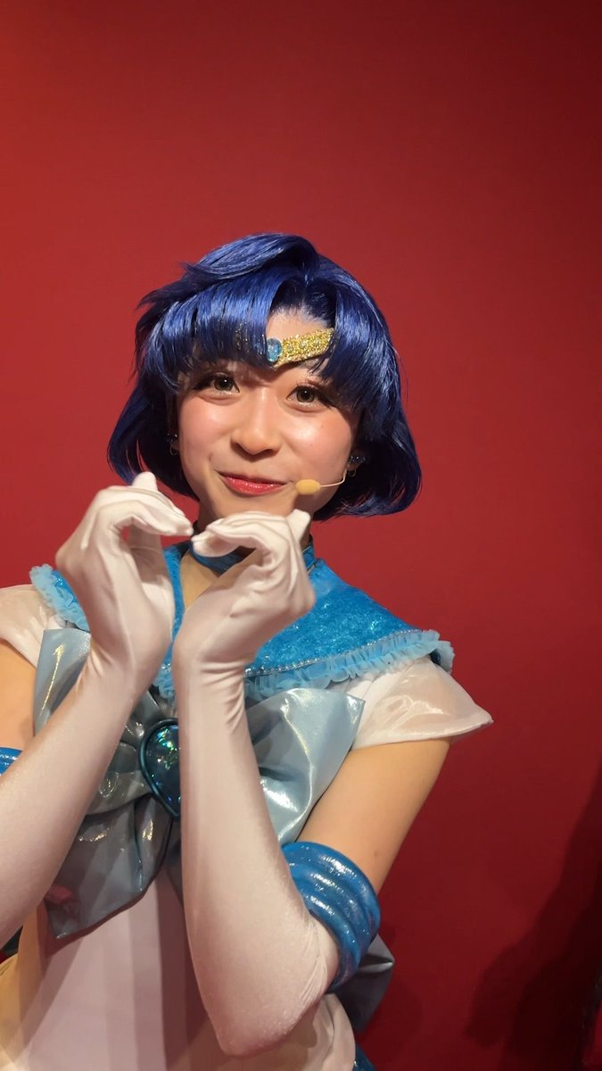 A young woman cosplaying as Sailor Mercury with blue hair and a gold tiara poses in a blue and white sailor outfit with white gloves against a red background.