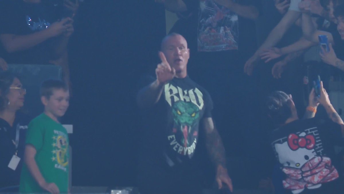 A bald man wearing an RKO 'EveryEverybody' t-shirt stands at a concert/event making an obscene gesture with his middle finger raised, while a young boy in a green shirt and audience members stand nearby.