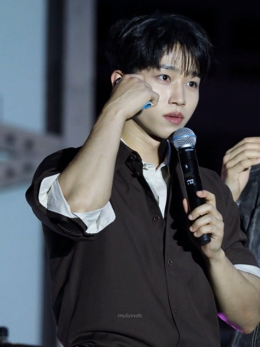 A young man with dark hair holding a Shure microphone while wearing a brown button-up shirt over a white top.
