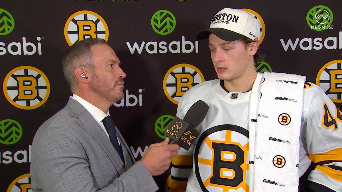 A reporter from NESN 360 holds a microphone up to Boston Bruins player James Hagens, who is wearing a #44 jersey and a Bruins cap with a Gillette Labs towel draped over his shoulder, during a postgame interview against a backdrop of Bruins and Wasabi logos. The image captures the moment after Hagens scored his first career NHL goal, as reflected in his comment about how it feels great.