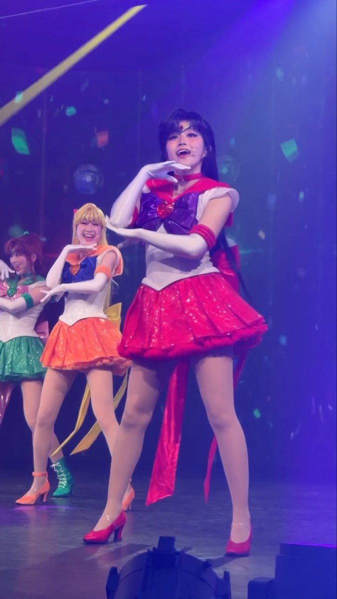 Performers in Sailor Moon-inspired costumes dance on a stage, with the central figure wearing a red and white outfit, all striking synchronized poses.