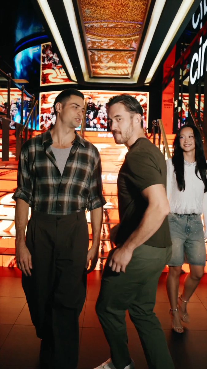 Three people posing for a photo in a brightly lit venue with red walls and large illuminated displays, with one person wearing a plaid shirt and another in a brown t-shirt.