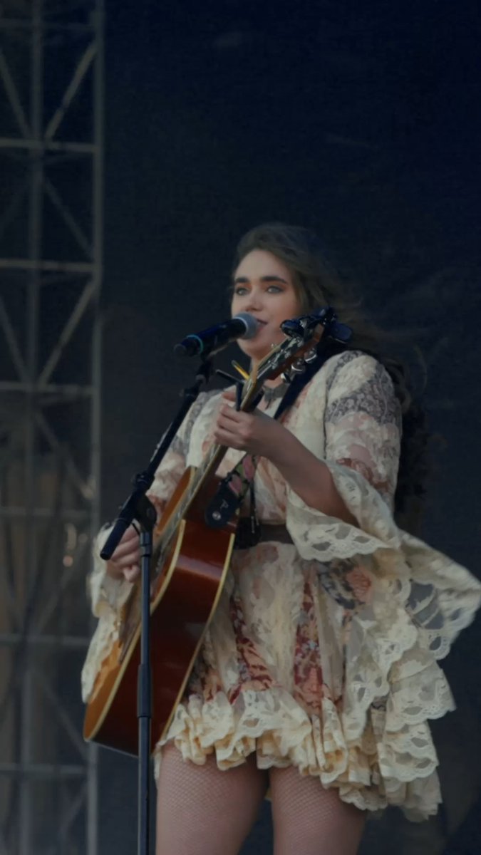 A young woman with long wavy brown hair performing on stage, wearing a cream-colored floral lace dress and singing into a microphone while playing an acoustic guitar.
