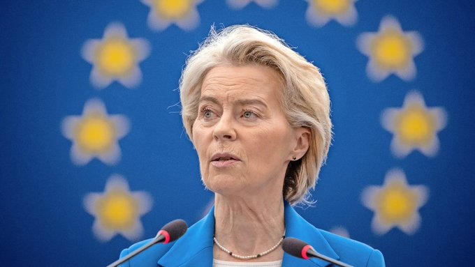 A blonde woman in a blue blazer speaks at a podium with microphones, standing before the blue and yellow star-patterned backdrop of the European Union flag.