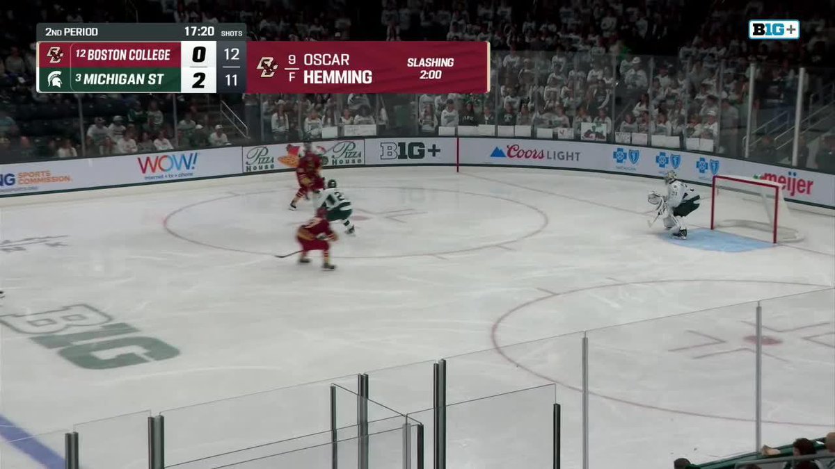 A hockey game is in progress on a Big+ broadcast showing Boston College trailing Michigan State 0-2 with 17:20 left in the 2nd period and 11:12 shots on goal, as a Michigan State player in green and white jerseys skates toward the Boston College goaltender. A penalty notification at the top indicates Oscar Hemming has a 2:00 slashing penalty. The caption's reference to a "strong kick save by Ravensbergen" aligns with the visible goaltender positioned in the crease appearing ready to make a save.