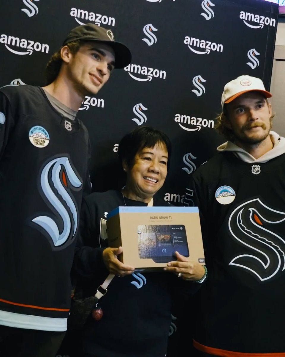 A woman holds an Echo Show 11 box while posing between two Seattle Kraken hockey players in branded jerseys in front of an Amazon and Kraken logo backdrop, illustrating the team's Prime Big Deal Days promotion with Amazon.