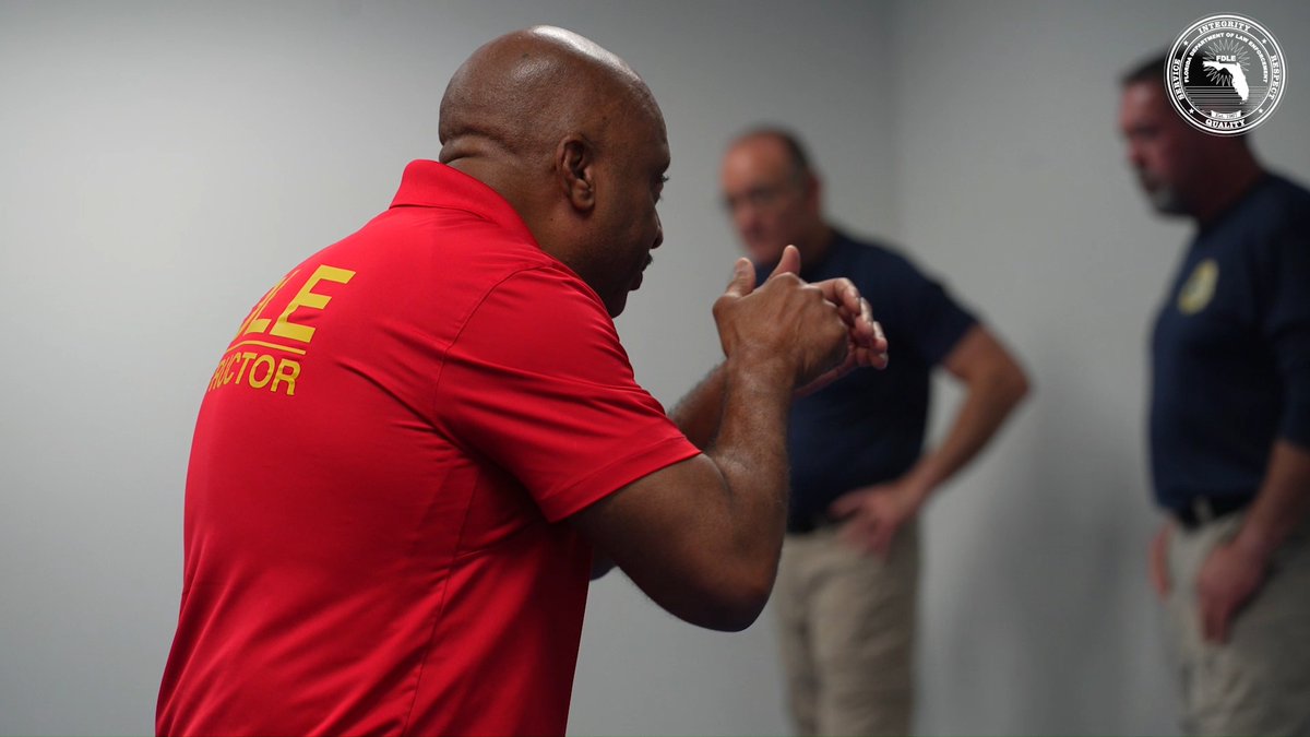 A bald Black man wearing a red 'FIRE INSTRUCTOR' polo shirt demonstrates hand positioning, with two other men in dark uniforms observing him in a training room.