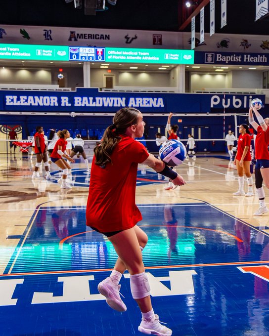 A female volleyball player in a red jersey serving a ball during a game at Eleanor R. Baldwin Arena, with the scoreboard visible in the background showing American Athletic Conference branding.