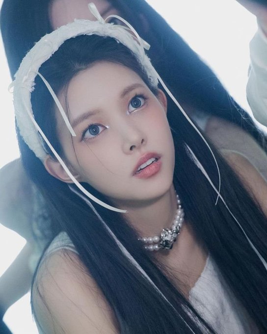 A close-up portrait of a young woman with long dark hair wearing a white lace headband with ribbons, a pearl choker necklace with a decorative brooch, and soft makeup.