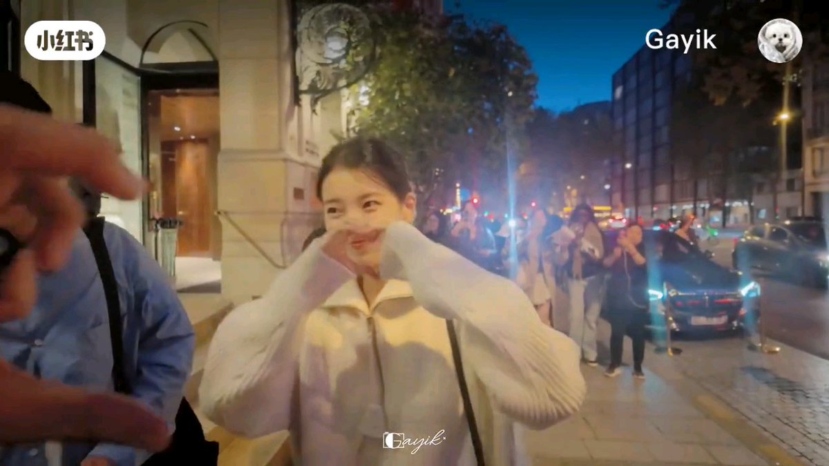 A young woman poses on a city street at dusk with a crowd behind her; the image is watermarked with '小红书' and 'Gayik'.