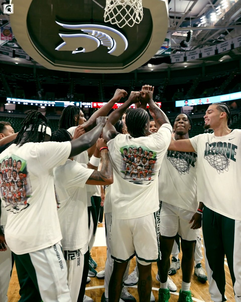 A basketball team in white Michigan State Spartans gear huddles under the hoop with their hands joined in a team gesture inside an arena.