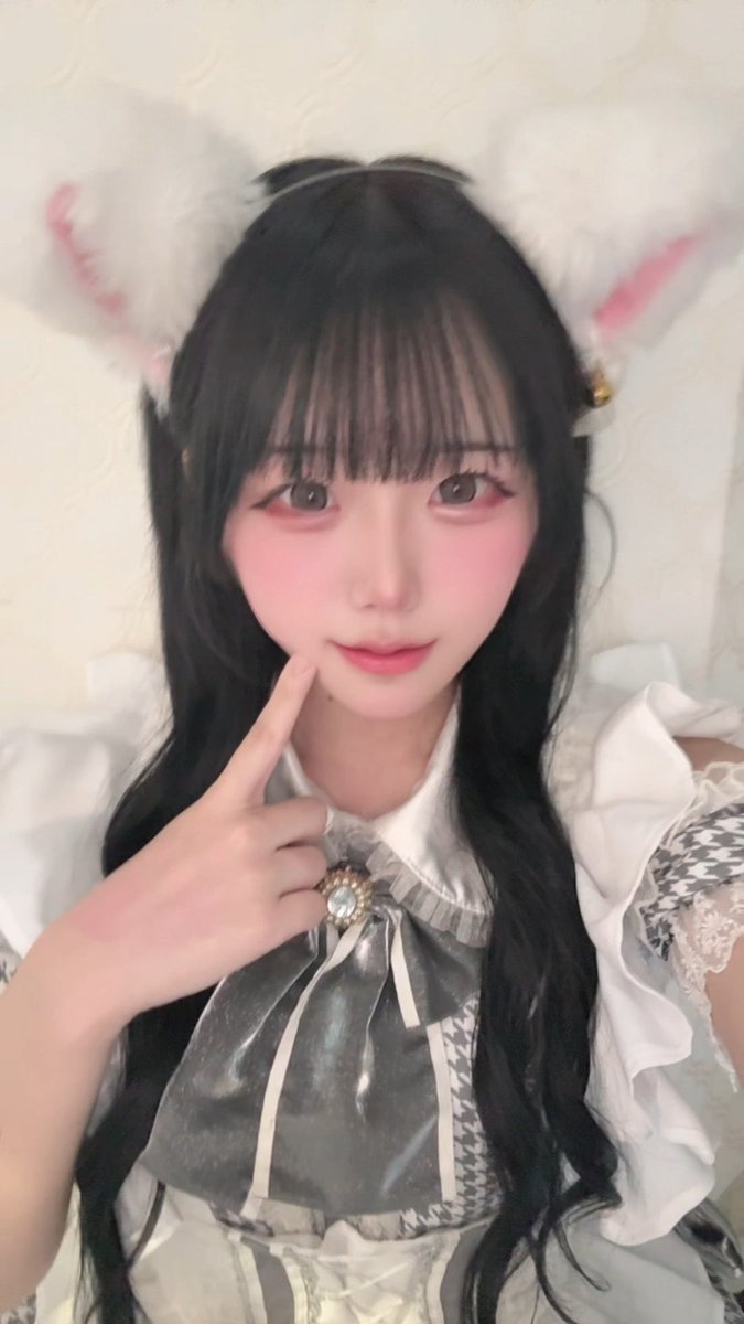 A young woman with long dark hair and cat-ear headwear poses for a selfie with her finger touching her cheek, wearing dramatic pink eye makeup.