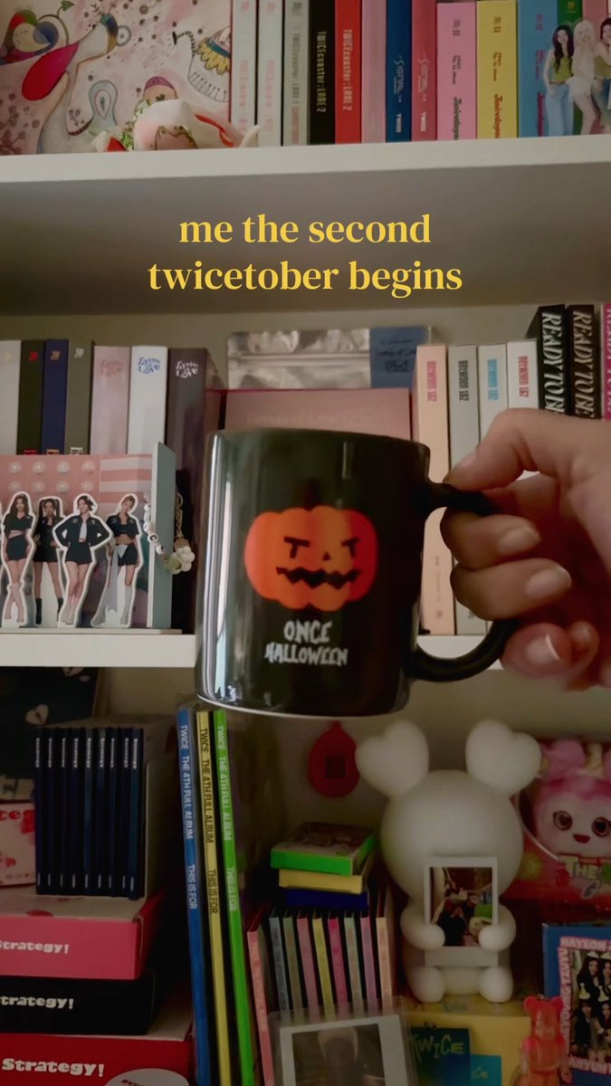 A hand holds up a black mug with a jack-o'-lantern design that says "ONCE HALLOWEEN," with overlay text "me the second twictober begins" against a background of K-pop merchandise shelves.