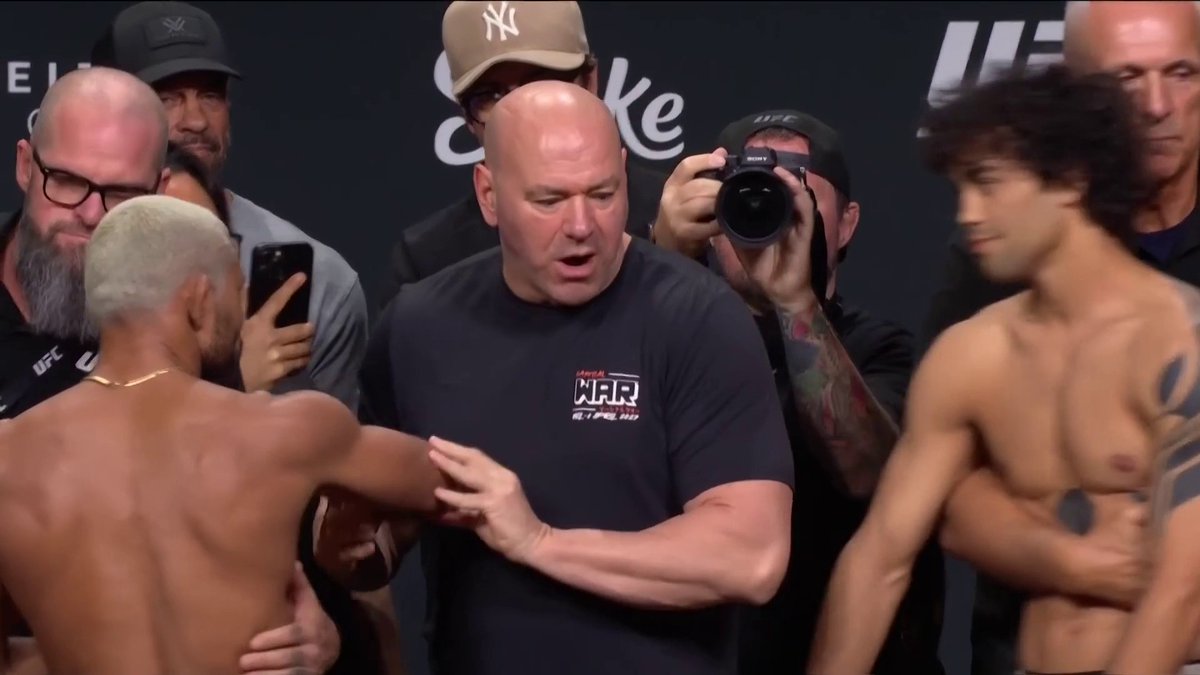 Dana White (wearing his "SMOKE WAR" shirt) separates two shirtless fighters during an intense final face-off at the UFC weigh-in event. The scene captures the tense moment between Figgy and Payton Talbott as they square off before their bout, surrounded by photographers and team members.